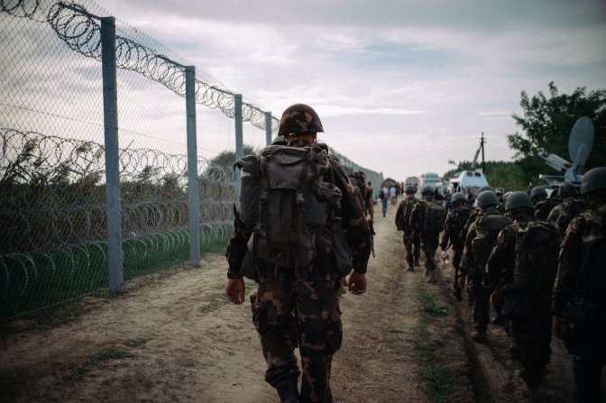 Une soldat lourdement équipé longe une frontière, hérissée de barbelés, 